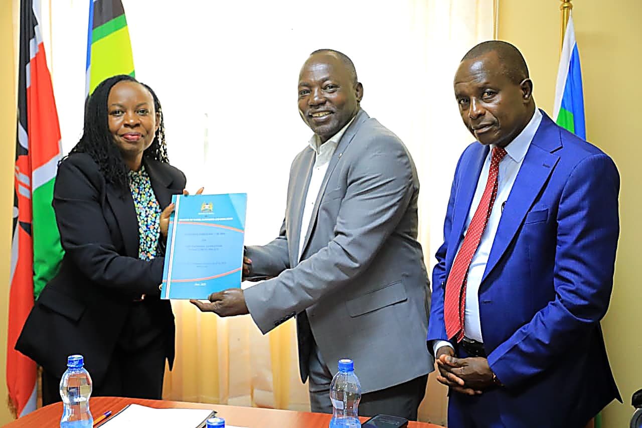 A document being handed over during a meeting, flanked by national and county flags.